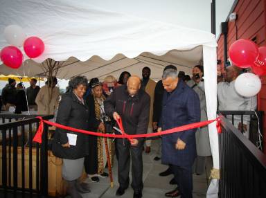 A woman and two men cutting a red ribbon with large scissors at an outdoor ribbon-cutting ceremony.