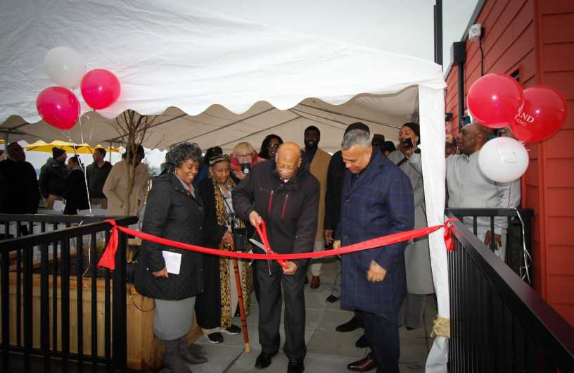 A woman and two men cutting a red ribbon with large scissors at an outdoor ribbon-cutting ceremony.